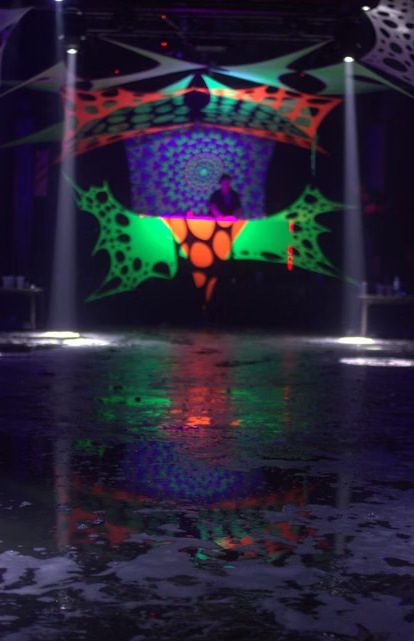 Estructura escénica con telas fluorescentes reactivas a luz UV, iluminada en verde y naranja.