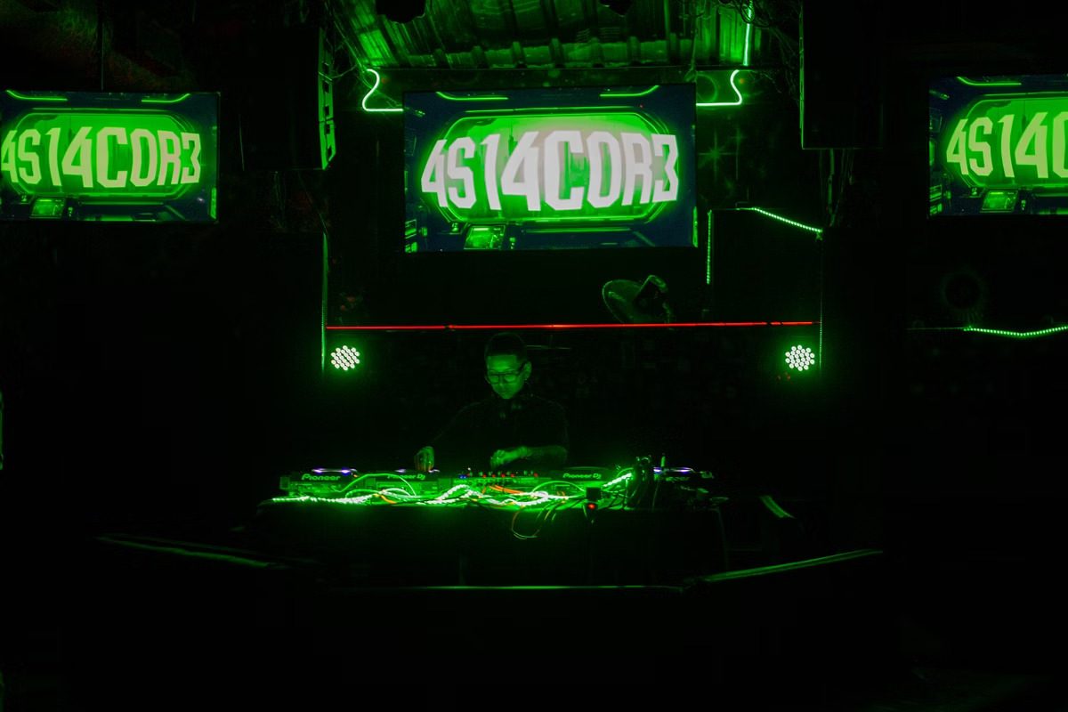 Cabina bajo pantallas verdes con el texto 4S14C0R3 y fixtures de par LED.
